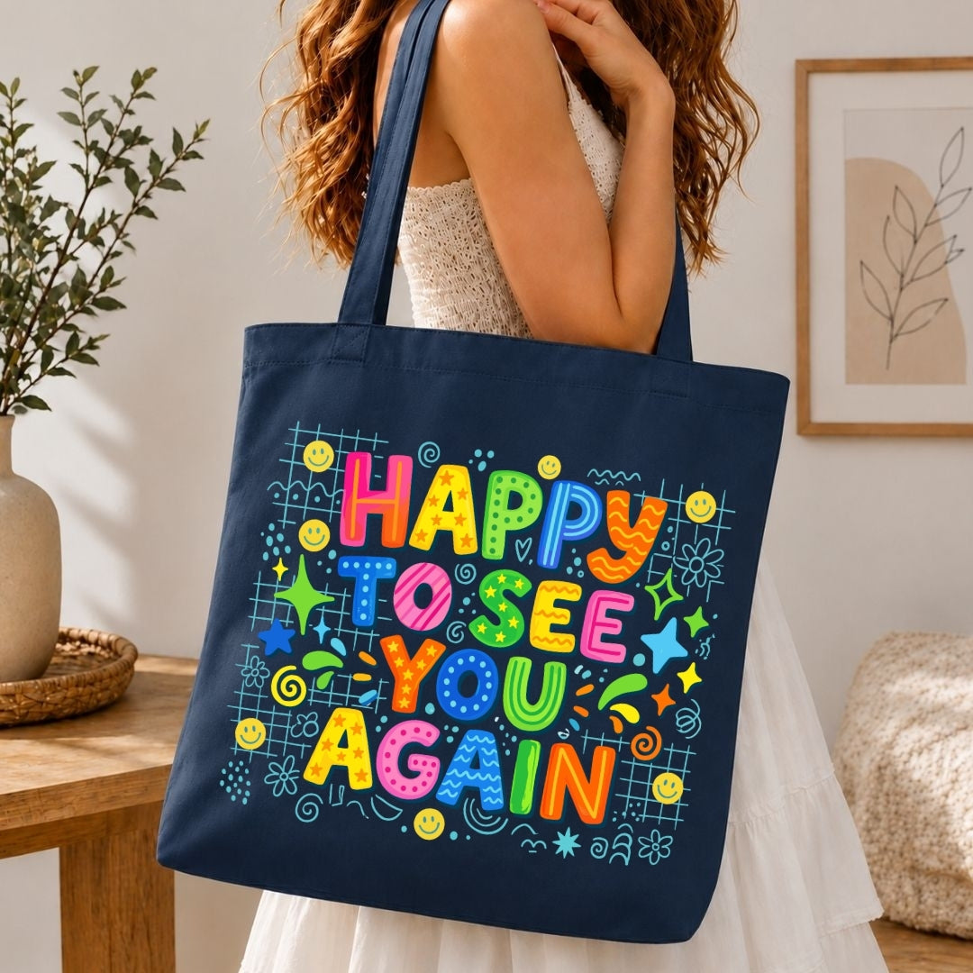 A woman holds a navy blue canvas tote bag with a colorful 'Happy to See You Again' graphic print, set in a warm, modern indoor room.