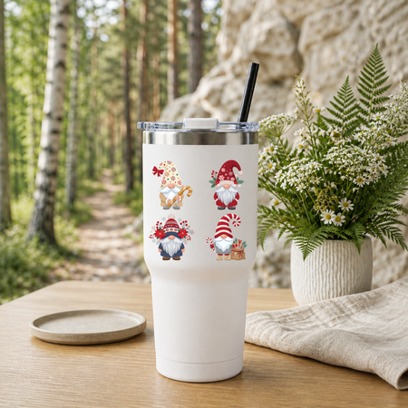 White travel tumbler with a black straw featuring a Christmas gnomes design, placed outdoors near a wooded trail.