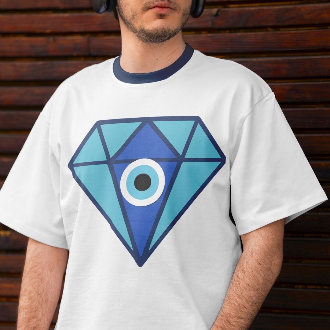 A man wearing a white ringer t-shirt with a navy blue crew neck collar and a large blue geometric diamond graphic with an eye design on the front, set against a dark brown wooden slat wall.