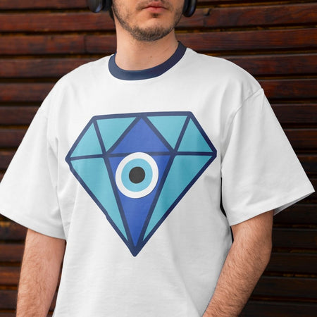 A man wearing a white ringer t-shirt with a navy blue crew neck collar and a large blue geometric diamond graphic with an eye design on the front, set against a dark brown wooden slat wall.