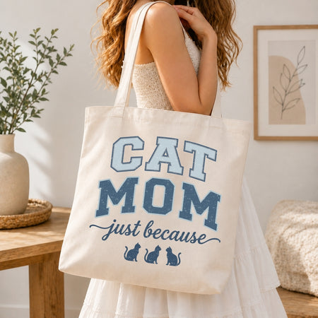 Woman in a white tiered dress carrying a canvas tote bag printed with a blue retro "Cat Mom just because" graphic over her shoulder.