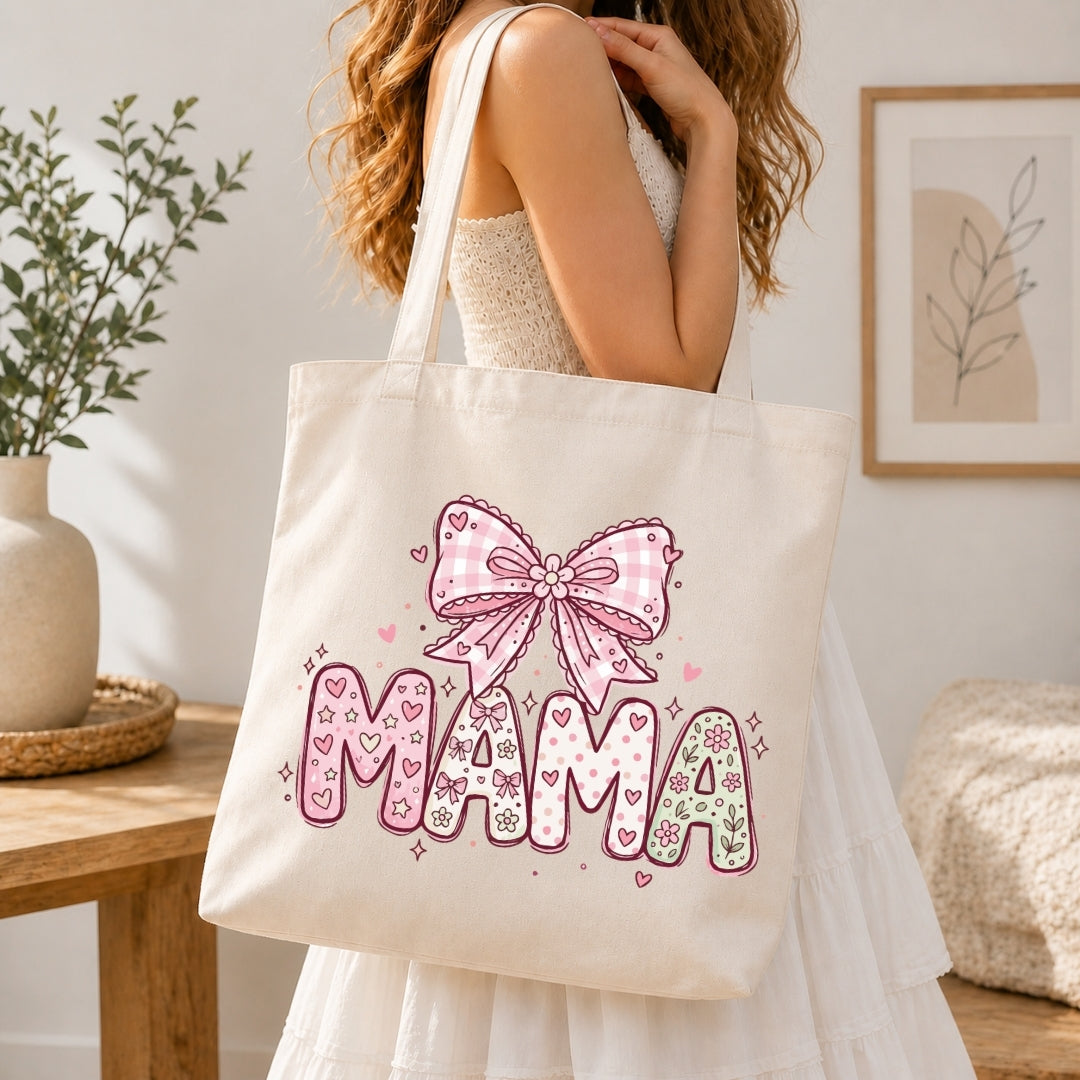 Woman in a white tiered dress carrying a cream canvas tote bag printed with a large pastel pink "MAMA" coquette gingham bow design over her shoulder.