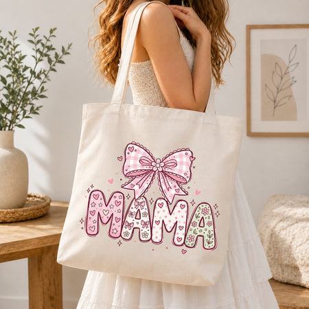 Woman in a white tiered dress carrying a cream canvas tote bag printed with a large pastel pink "MAMA" coquette gingham bow design over her shoulder.