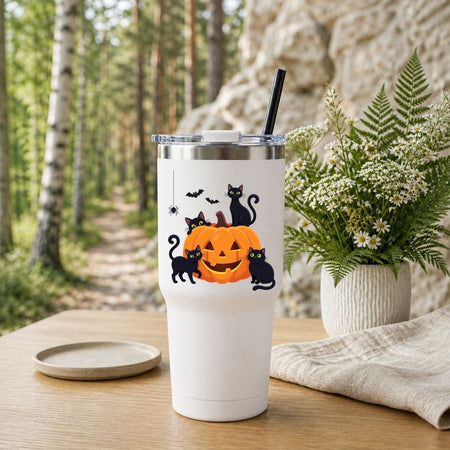 White travel tumbler with a black straw featuring a cute Halloween black cats and pumpkin graphic, set outdoors on a wooden table with a forest background