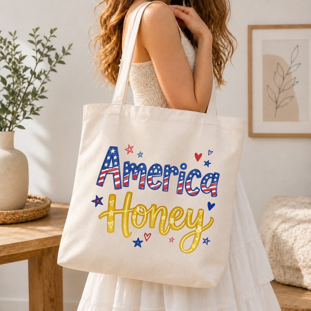 A woman in a white dress carries a cream canvas tote bag with 'America Honey' written in red, white, and blue patriotic lettering with yellow script and small red, white, and blue star and heart accents. The scene is set in a bright, modern room with a wooden table, potted plant, and framed wall art.