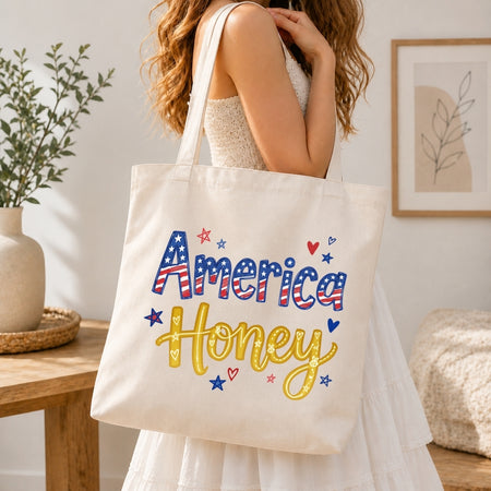 A woman in a white dress carries a cream canvas tote bag with 'America Honey' written in red, white, and blue patriotic lettering with yellow script and small red, white, and blue star and heart accents. The scene is set in a bright, modern room with a wooden table, potted plant, and framed wall art.