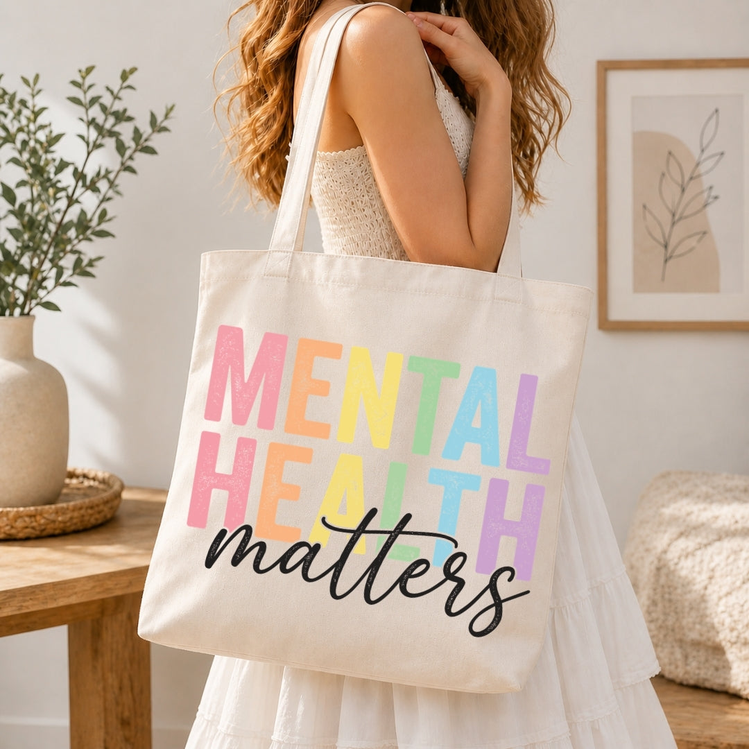 A woman in a white dress carries a cream canvas tote bag with the words 'MENTAL HEALTH matters' printed in pastel rainbow letters and black script. The scene is set in a bright, modern room with a wooden table, potted plant, and framed wall art.