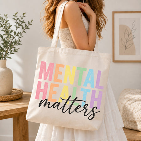 A woman in a white dress carries a cream canvas tote bag with the words 'MENTAL HEALTH matters' printed in pastel rainbow letters and black script. The scene is set in a bright, modern room with a wooden table, potted plant, and framed wall art.