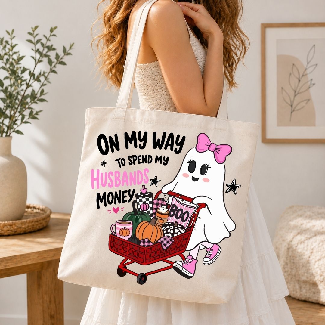A person carrying a natural canvas tote bag printed with the ghost pushing a shopping cart and the "On My Way to Spend My Husbands Money" graphic.