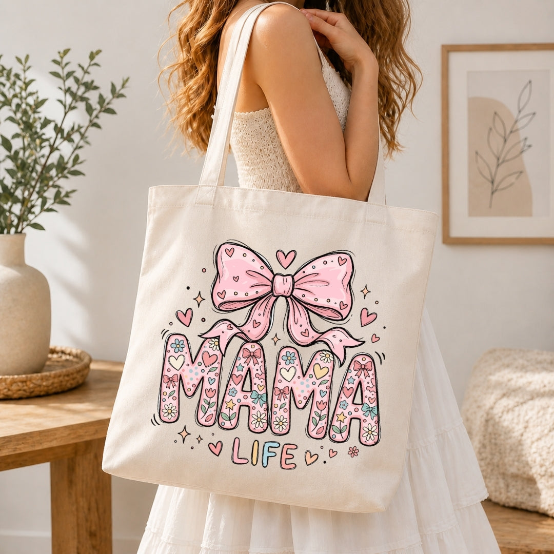 Woman in a white dress carrying a cream canvas tote bag printed with a large pastel floral "MAMA LIFE" coquette illustration over her shoulder.