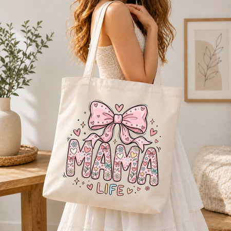 Woman in a white dress carrying a cream canvas tote bag printed with a large pastel floral "MAMA LIFE" coquette illustration over her shoulder.