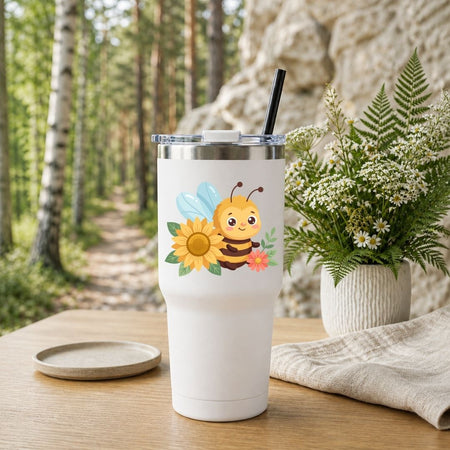 A white insulated tumbler with a clear lid and black straw, featuring a cute cartoon bee, sunflower, and flower graphic, set on a wooden table in a forest path scene with a potted fern in the background.