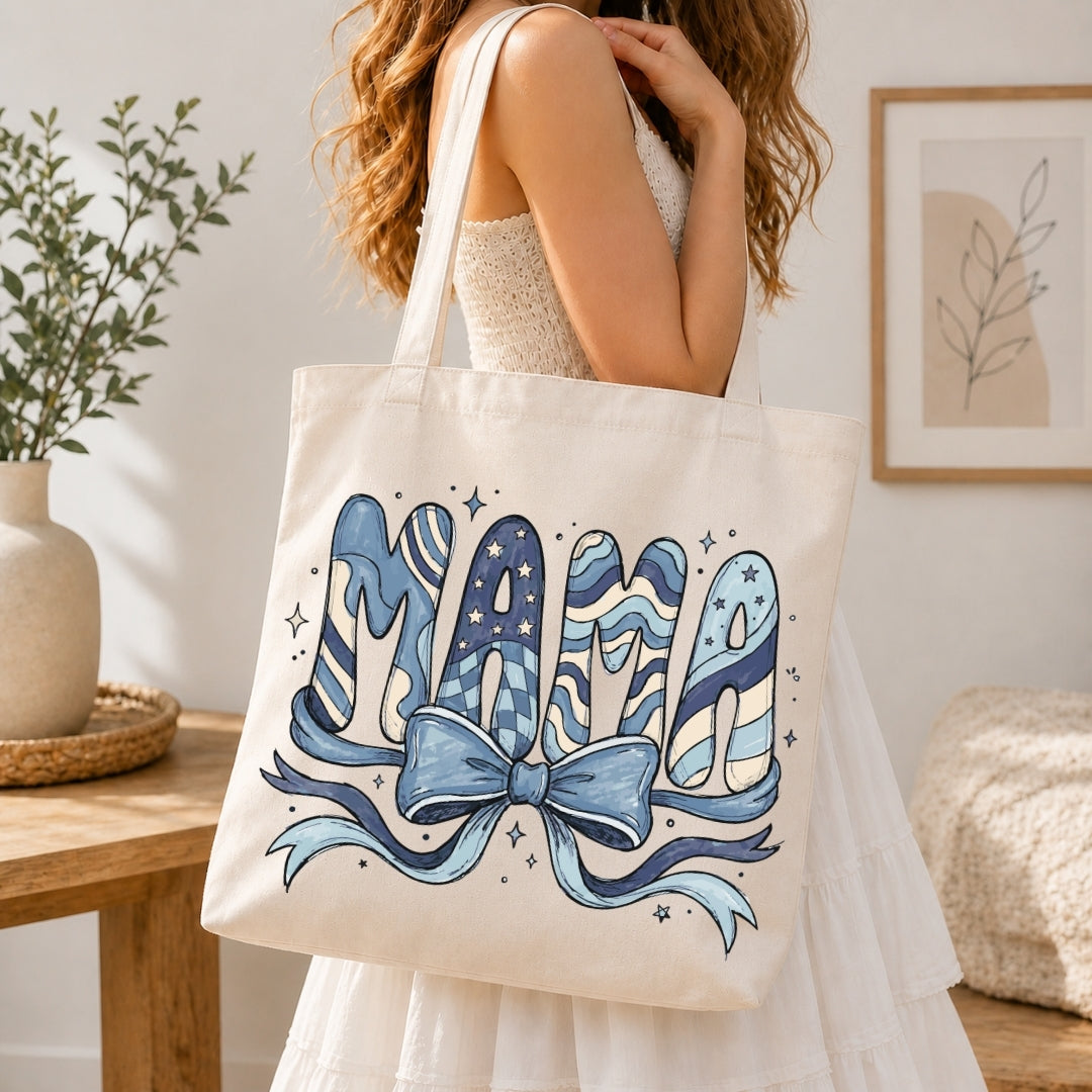 Woman in a white tiered dress carrying a cream canvas tote bag printed with a large blue "MAMA" coquette pattern ribbon design over her shoulder.
