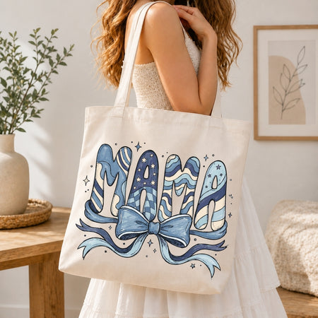 Woman in a white tiered dress carrying a cream canvas tote bag printed with a large blue "MAMA" coquette pattern ribbon design over her shoulder.