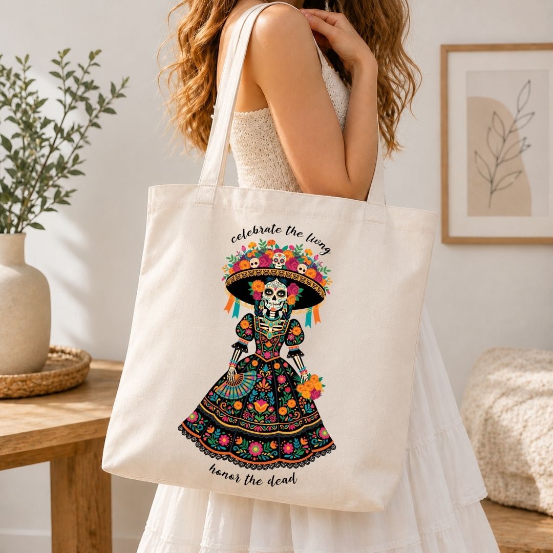 A woman in a white dress carries a cream canvas tote bag featuring a colorful Dia de los Muertos sugar skull illustration with the text 'celebrate the living' and 'remember the dead'.