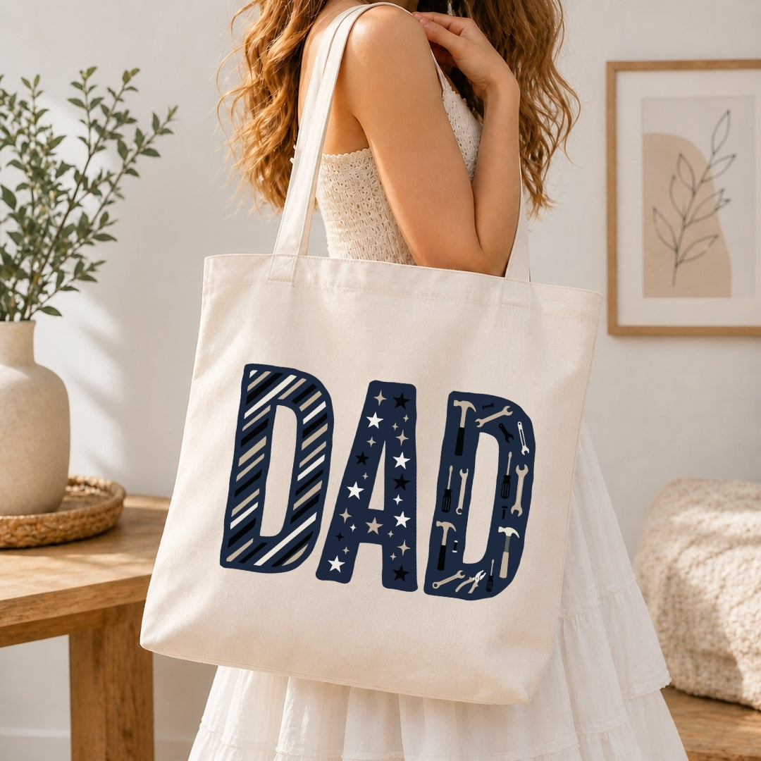 A woman in a white dress carries a cream canvas tote bag with the word 'DAD' printed in large navy letters filled with small white star and stripe patterns, set in a bright, modern room.