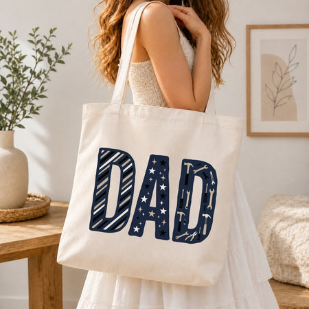 A woman in a white dress carries a cream canvas tote bag with the word 'DAD' printed in large navy letters filled with small white star and stripe patterns, set in a bright, modern room.