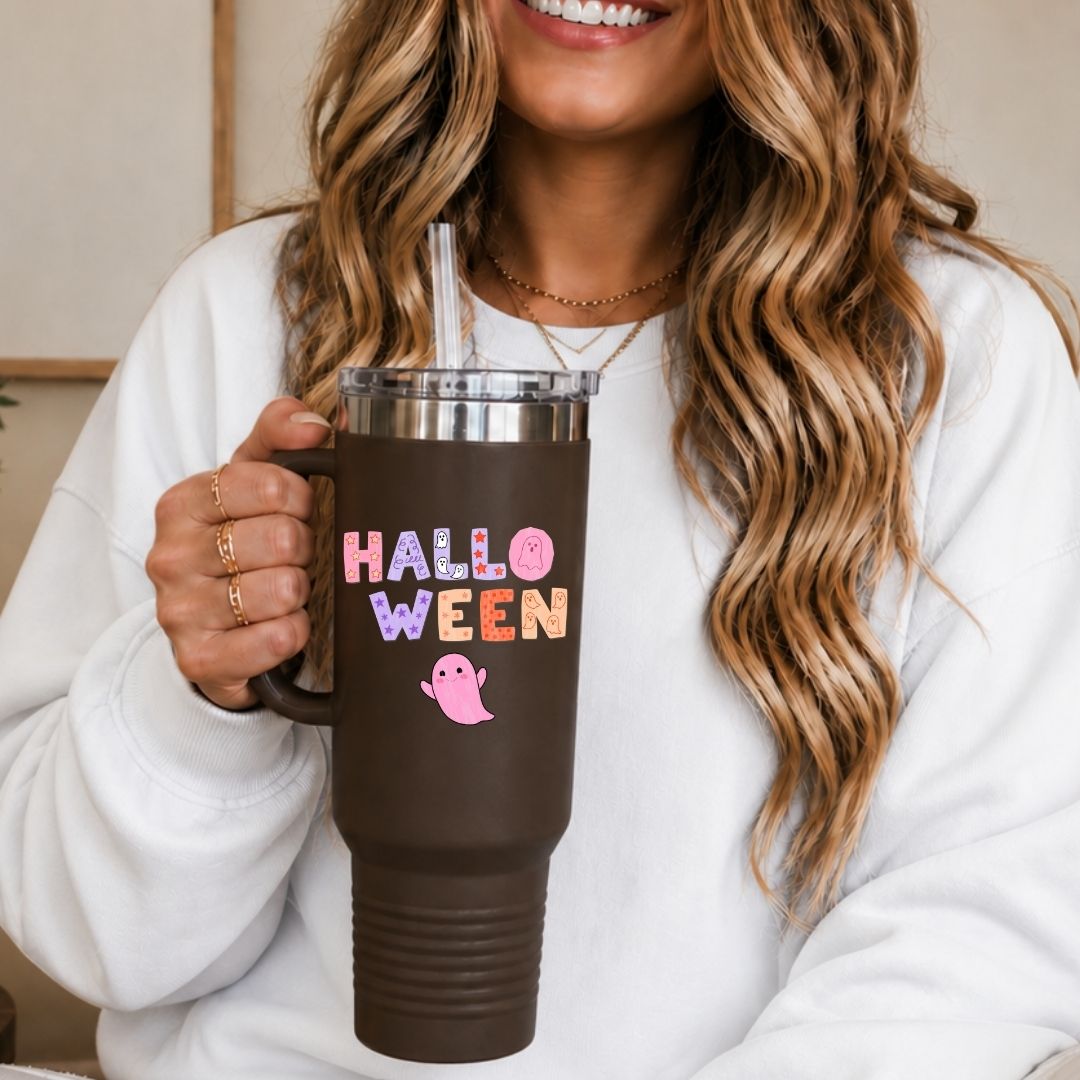 A woman in a white sweatshirt holds a dark brown insulated tumbler with a clear lid and straw. The tumbler has a metallic silver band near the top and colorful 'HALLOWEEN' text with a pink ghost graphic on the front.
