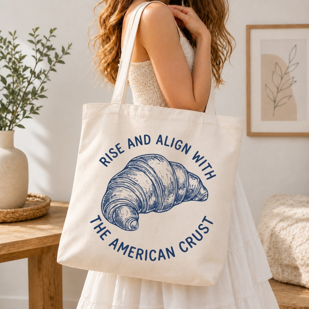 Woman in a white tiered dress carrying a cream-colored canvas tote bag printed with a large blue retro croissant sketch over her shoulder.