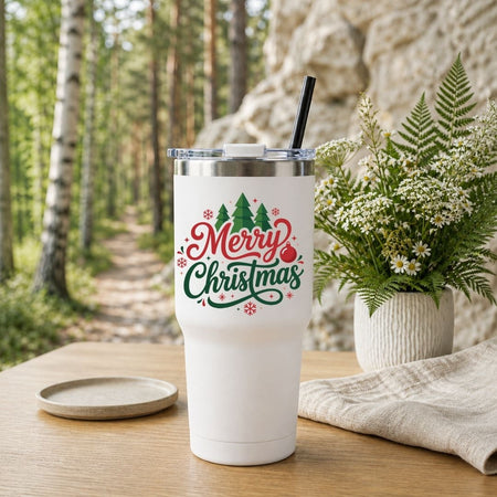 White travel tumbler with a black straw featuring a "Merry Christmas" holiday graphic, set outdoors on a wooden table with a forest background.