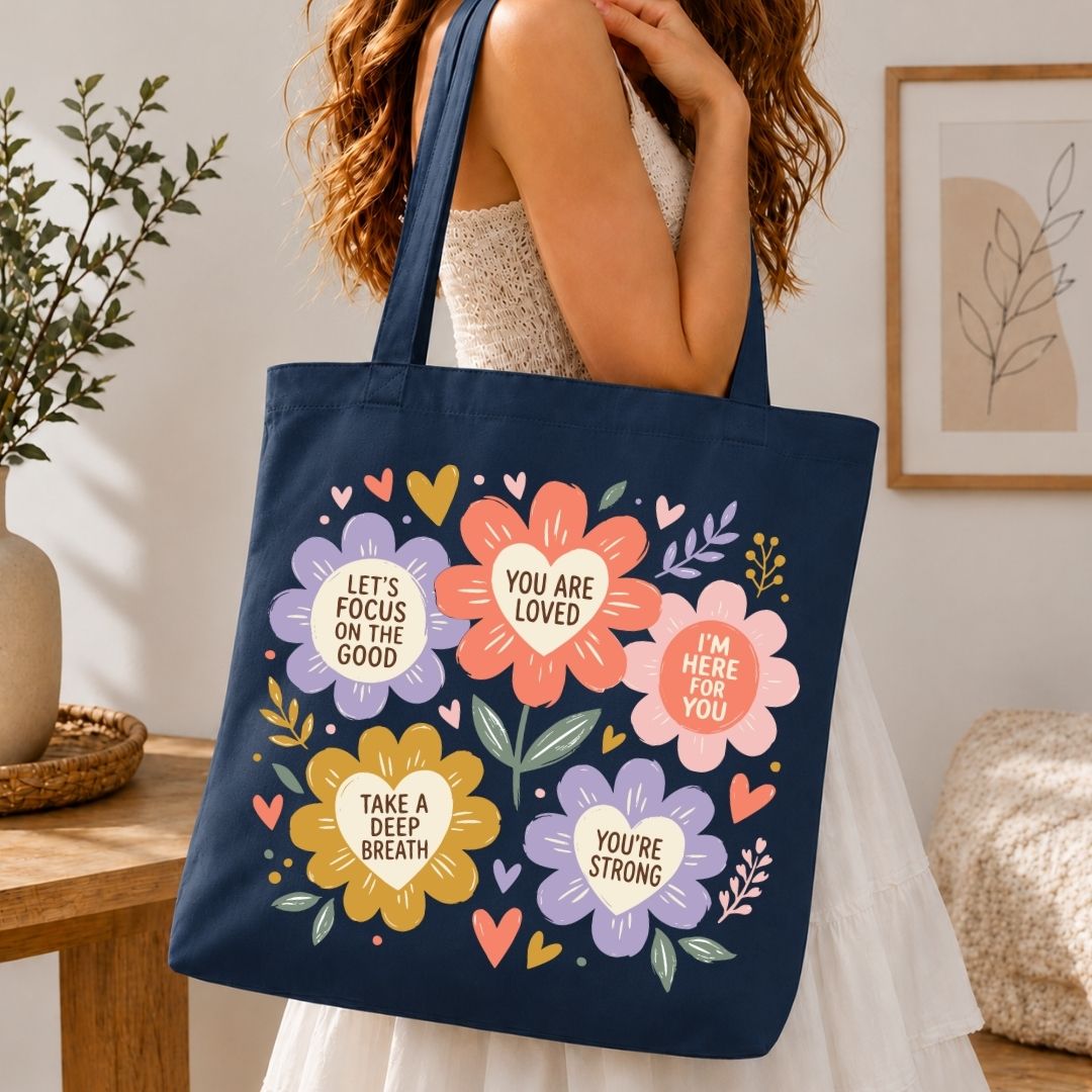 A navy blue canvas tote bag with colorful floral graphics and positive text quotes, carried by a woman in a white dress in a cozy indoor setting.