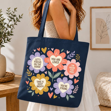A navy blue canvas tote bag with colorful floral graphics and positive text quotes, carried by a woman in a white dress in a cozy indoor setting.