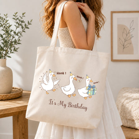 Person in a white dress carrying a canvas tote bag printed with the humorous "It's My Birthday" geese graphic.