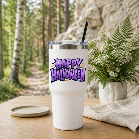 White travel tumbler with a black straw featuring a purple "Happy Halloween" design, placed outdoors near a nature path.