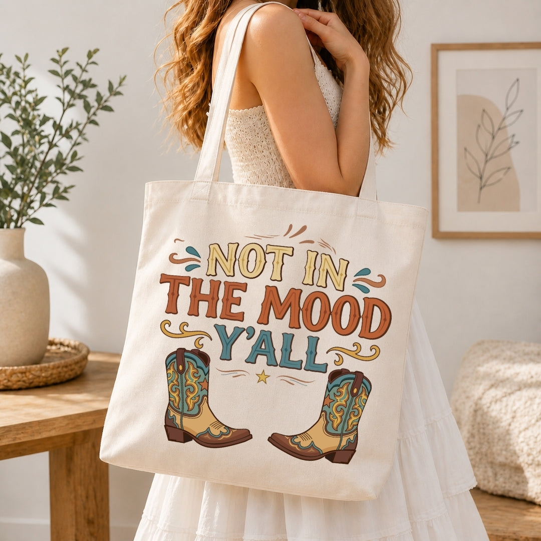 A woman in a white dress carries a cream canvas tote bag with a western-themed print that reads 'NOT IN THE MOOD Y'ALL' above a pair of colorful cowboy boots. The photo is taken in a bright, modern room with a wooden table, potted plant, and framed wall art.