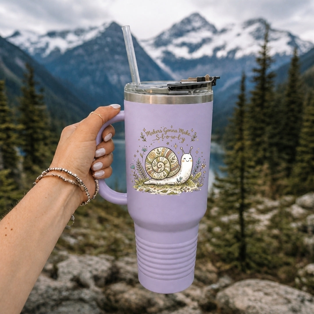 Hand holding a light purple insulated tumbler featuring the illustrated snail graphic, set outdoors against a scenic mountain and lake background.