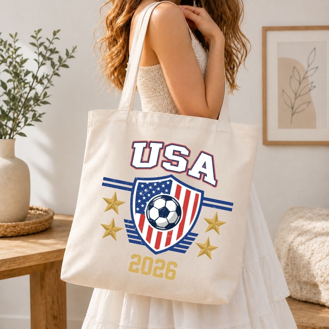 Person in a white dress carrying a canvas tote bag printed with the USA 2026 soccer graphic and gold stars.