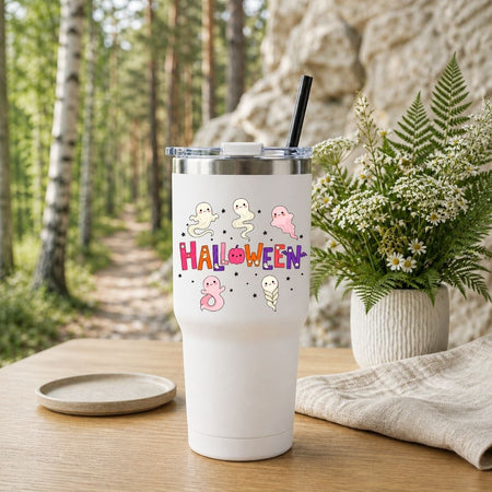 A white insulated tumbler with a clear lid and black straw, printed with cute cartoon ghosts and the word “HALLOWEEN” in colorful letters, set on a wooden table outdoors with a blurred forest and rock background, next to a potted fern and a small dish.