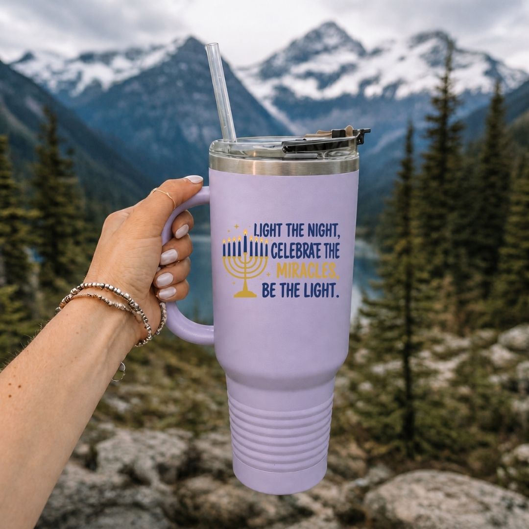 A lavender insulated tumbler with a clear straw is held by a hand in front of a mountain and forest background. The tumbler has a printed design with a menorah and the text, 'LIGHT THE NIGHT, CELEBRATE THE MIRACLES, BE THE LIGHT.