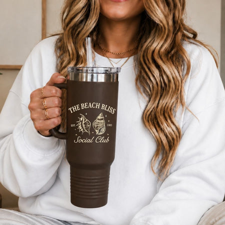 A woman in a white sweatshirt holding a 40oz brown travel tumbler with a handle, featuring a high quality cream sublimation print of a vintage seashell beach club design.