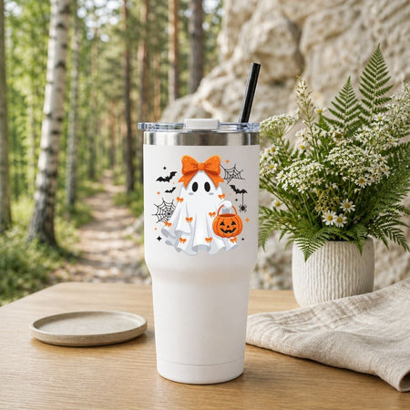 White travel tumbler with a black straw featuring a cute Halloween ghost graphic, set outdoors on a wooden table with a forest background.