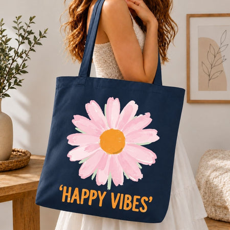 A navy blue canvas tote bag with a large pink daisy graphic and the text 'HAPPY VIBES' in orange, carried by a woman in a white dress in a bright indoor setting.