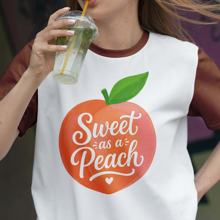 Person wearing a white t-shirt with a peach graphic and 'Sweet as a Peach' text, holding a drink.
