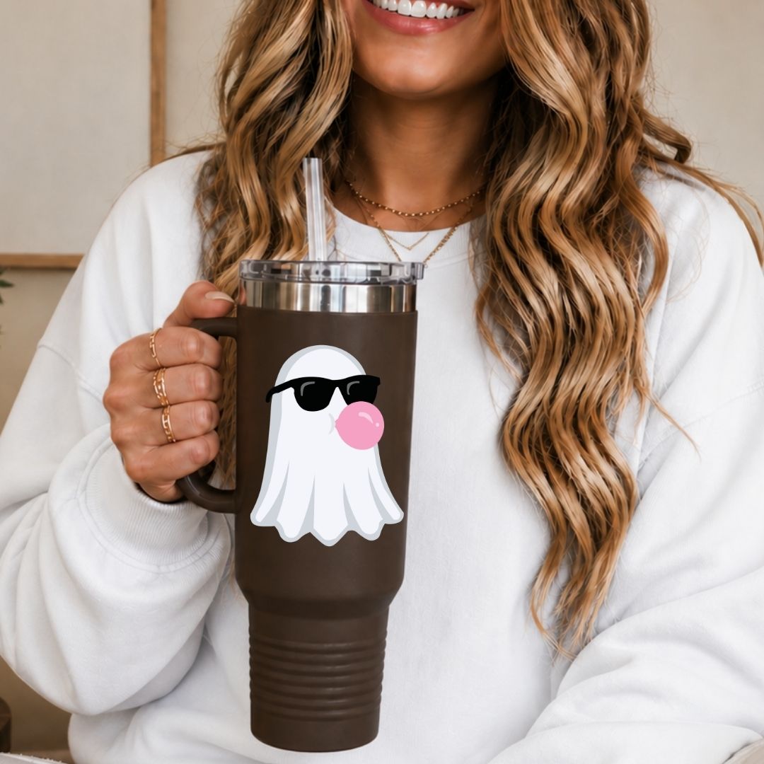 A woman in a white sweatshirt holds a brown insulated tumbler with a clear lid and straw. The tumbler has a white ghost graphic wearing black sunglasses and blowing a pink bubblegum bubble.