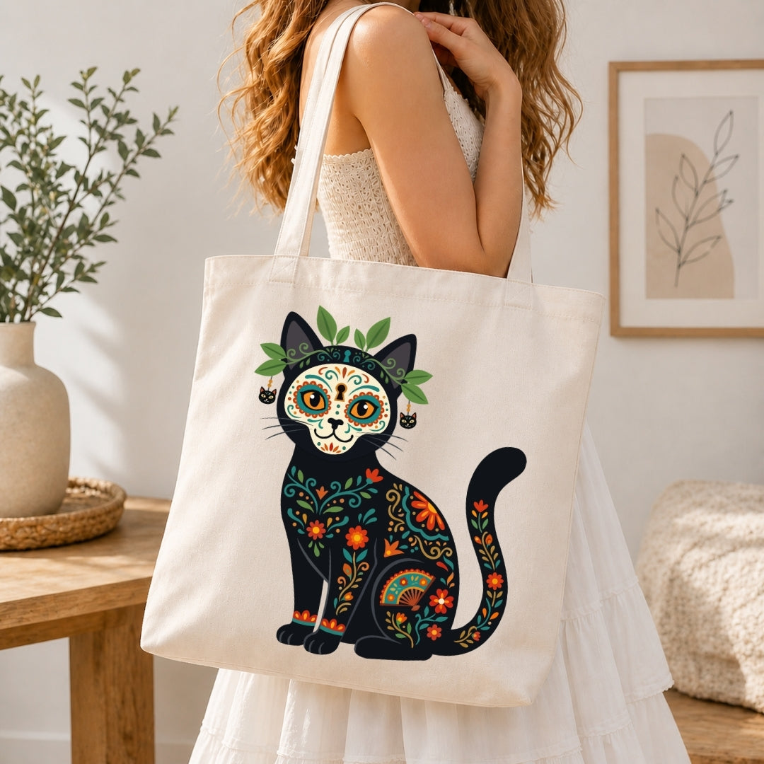 A woman in a white dress carries a cream canvas tote bag featuring a colorful Day of the Dead cat design. The background shows a softly lit, modern interior with a wooden table, a potted plant, and framed wall art.