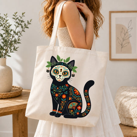 A woman in a white dress carries a cream canvas tote bag featuring a colorful Day of the Dead cat design. The background shows a softly lit, modern interior with a wooden table, a potted plant, and framed wall art.