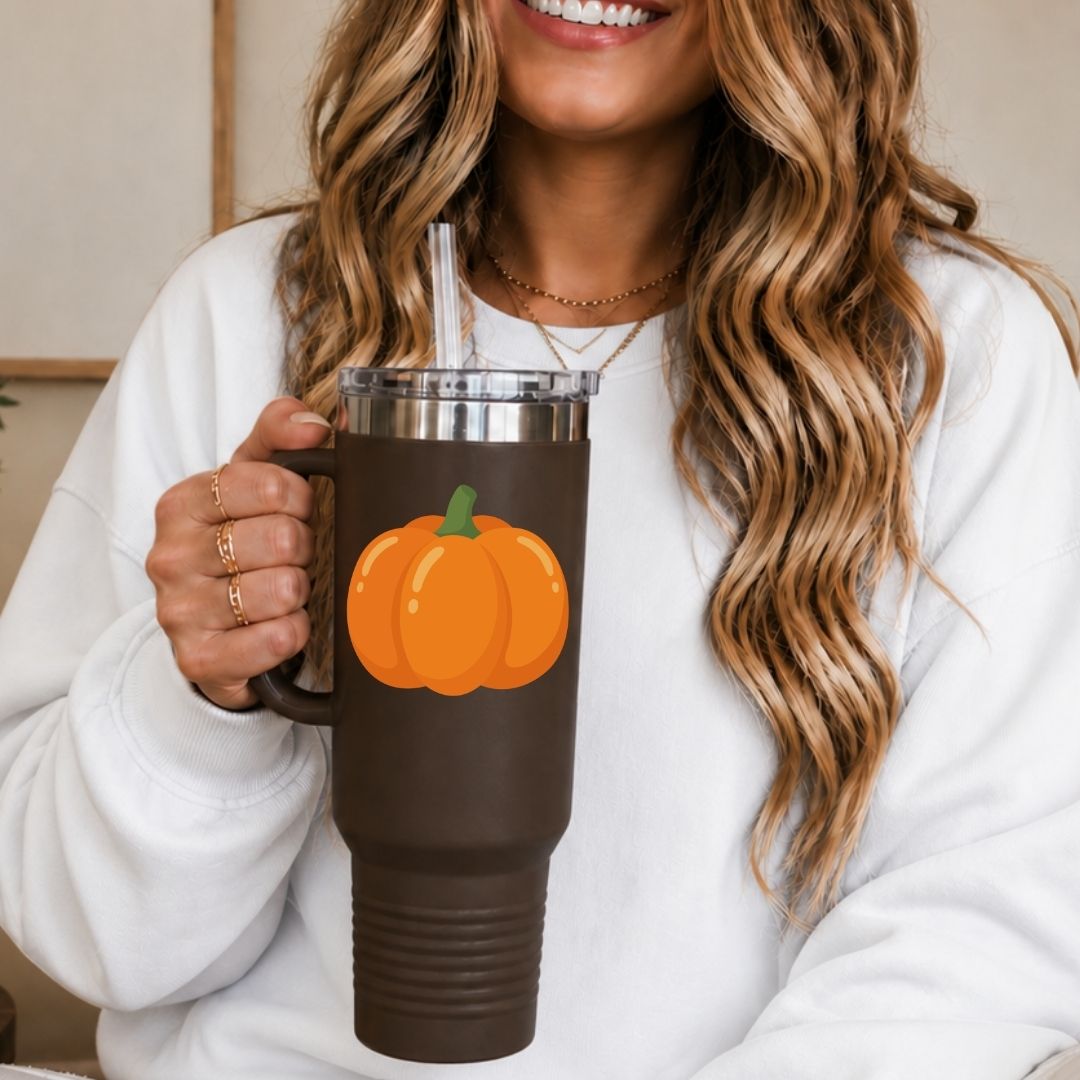 A woman in a white sweatshirt holds a dark brown insulated tumbler with a clear straw and a printed orange pumpkin graphic on the front.