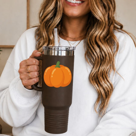 A woman in a white sweatshirt holds a dark brown insulated tumbler with a clear straw and a printed orange pumpkin graphic on the front.