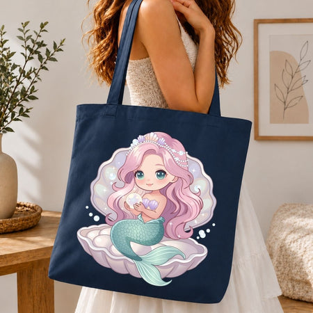 A navy blue canvas tote bag with a cute mermaid illustration, carried by a woman in a white dress in a cozy indoor setting.
