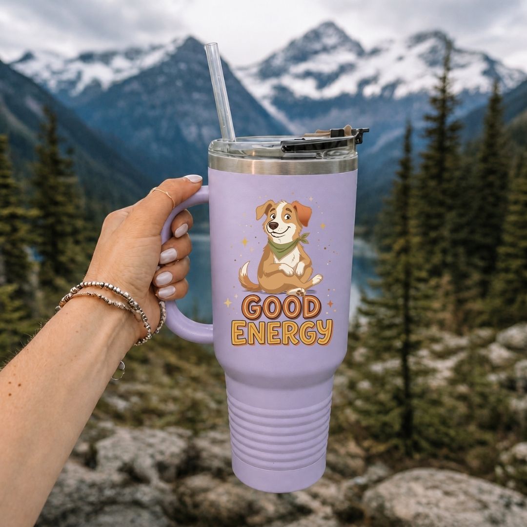 A hand holds a lavender insulated tumbler with a clear straw in front of a mountain and forest background. The tumbler features a cartoon dog wearing a green bandana and the words 'GOOD ENERGY' in orange.