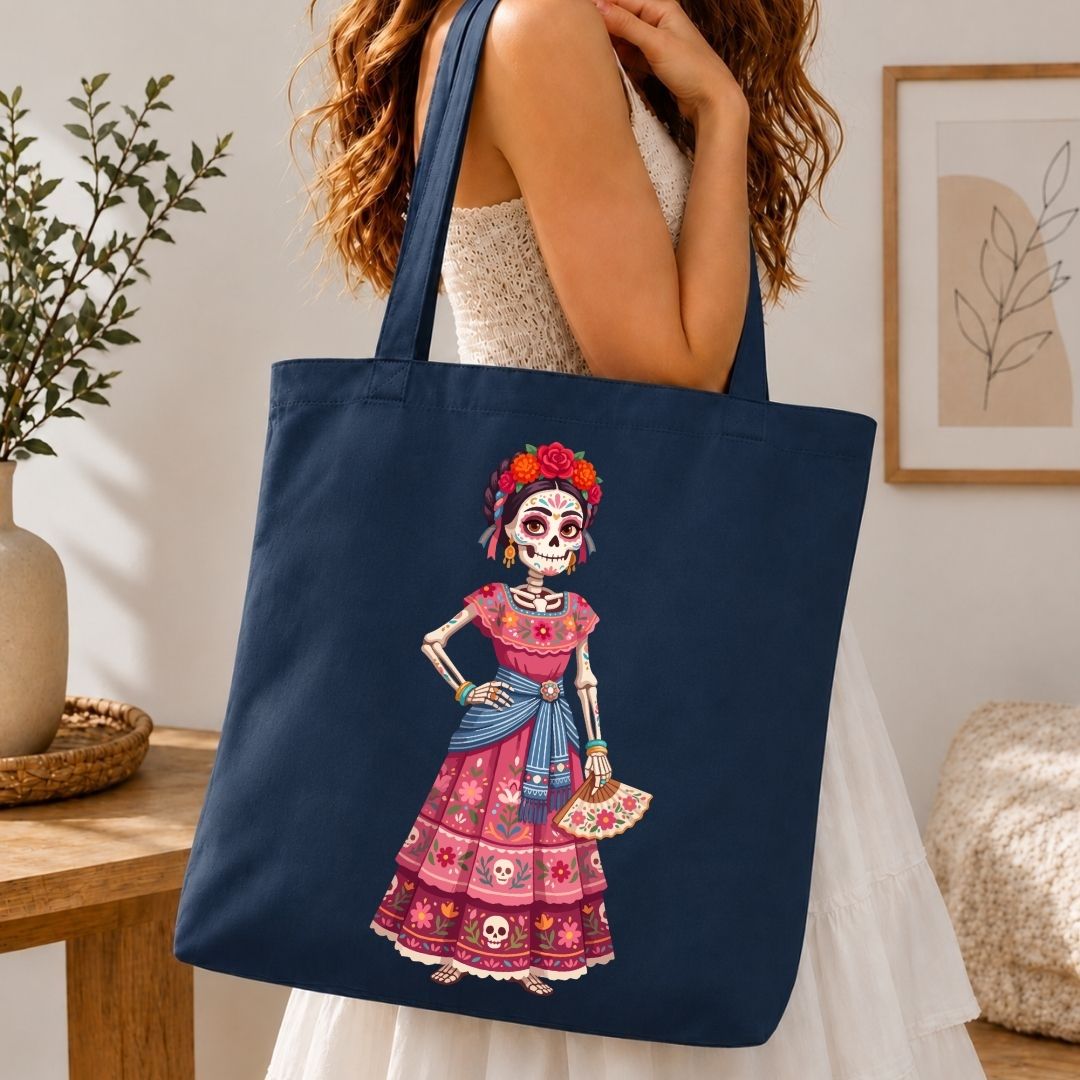 A navy blue canvas tote bag with a colorful Day of the Dead skeleton woman graphic, carried by a woman in a white dress in a warm, modern room setting.