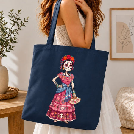 A navy blue canvas tote bag with a colorful Day of the Dead skeleton woman graphic, carried by a woman in a white dress in a warm, modern room setting.