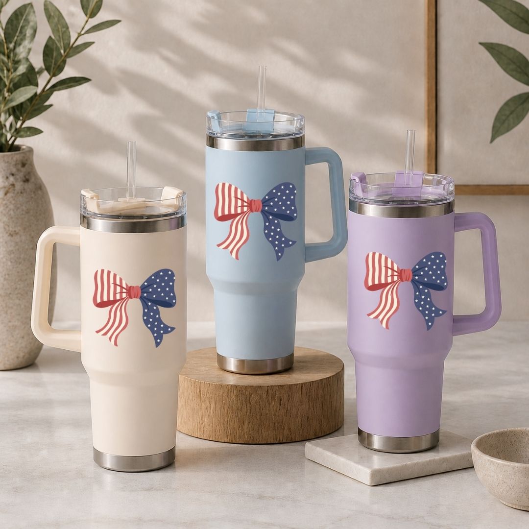 Three insulated travel tumblers in cream, light blue, and lavender purple arranged on a table, all showcasing the American flag ribbon bow design.