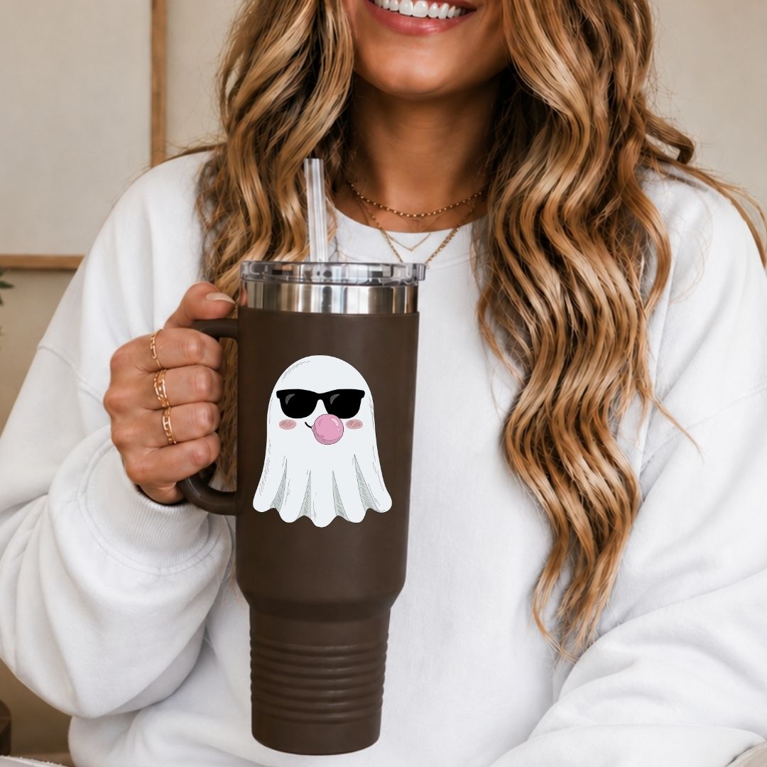 A woman in a white sweatshirt holds a dark brown insulated tumbler with a clear lid and straw. The tumbler has a cartoon ghost graphic wearing black sunglasses and a pink tongue sticking out.