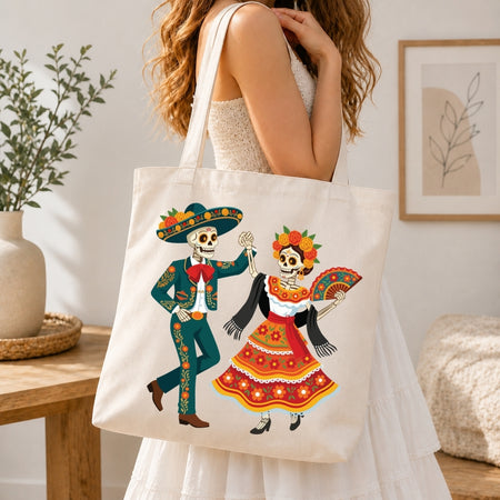 A woman in a white dress carries a cream canvas tote bag featuring a colorful Day of the Dead sugar skull illustration. The scene is set in a bright, modern room with a wooden table, potted plant, and framed wall art.