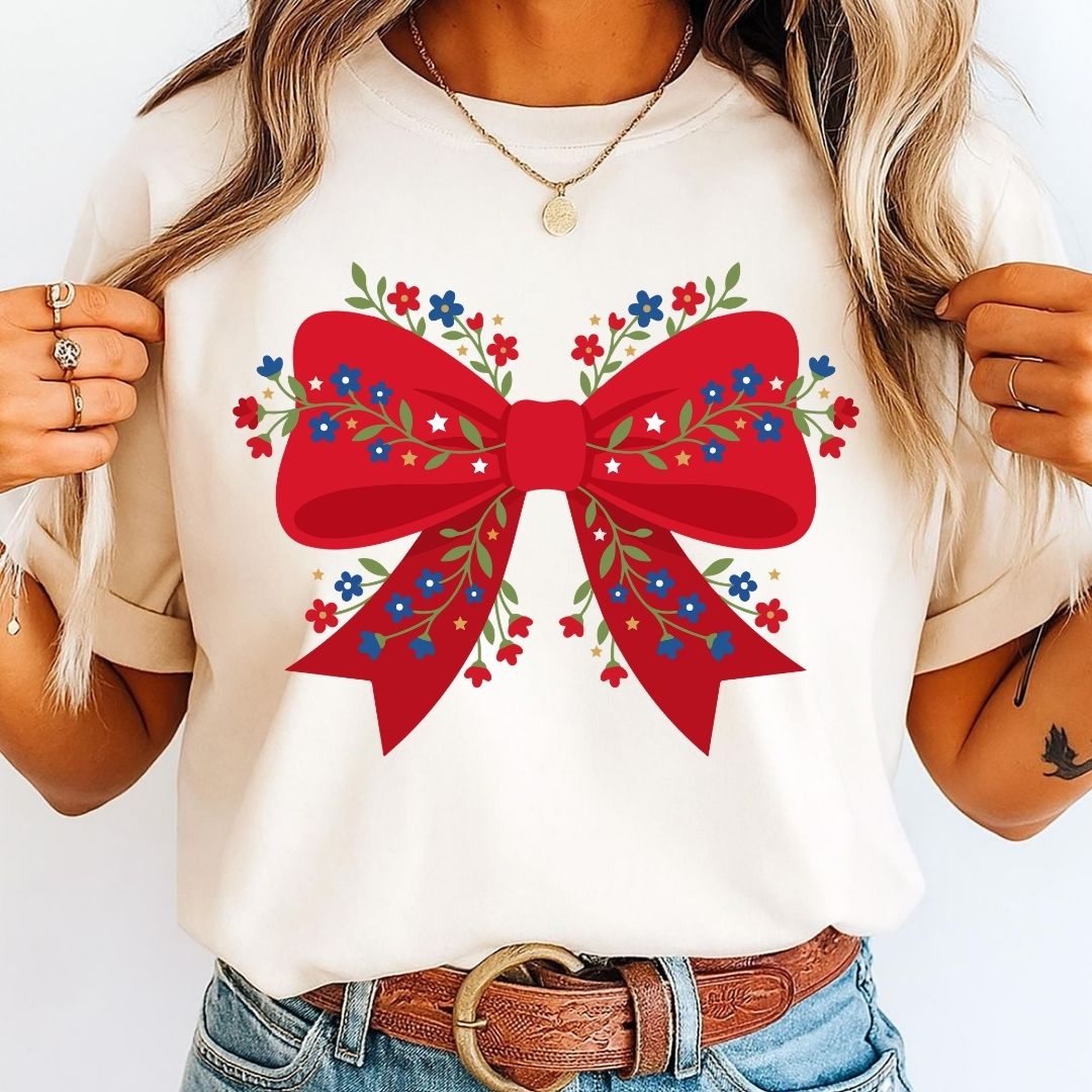 A woman wearing a cream-colored t-shirt with a large red bow and colorful floral embroidery graphic on the front, paired with blue jeans and a brown belt.